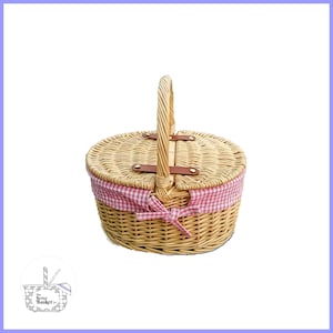 May include: A round wicker picnic basket with a pink and white checkered fabric lining and a brown leather handle. The basket has a hinged lid and a bow on the front.