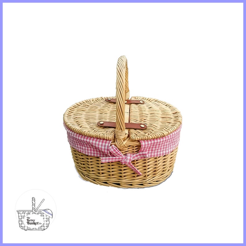 May include: A round wicker picnic basket with a pink and white checkered fabric lining and a brown leather handle. The basket has a hinged lid and a bow on the front.