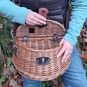 Op de afbeelding: Een bruine, ronde vismand van gevlochten wilgentenen met zwarte leren details en een metalen sluiting. De mand heeft een schouderriem en een kleine opening aan de bovenkant.