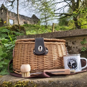 May include: A round woven wicker basket with a dark brown leather-like closure, a wooden scrubbing brush, and a white enamel mug featuring a black graphic illustration and the text "Some of us bloom in the gloom" printed on the side.