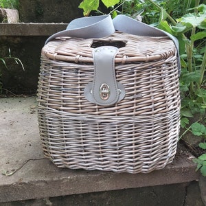 May include: A wicker basket with a leather handle and a gray fabric strap. The basket is woven with a light brown and white pattern.