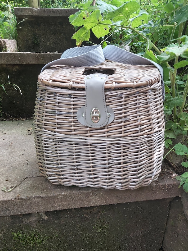 May include: A wicker basket with a leather handle and a gray fabric strap. The basket is woven with a light brown and white pattern.