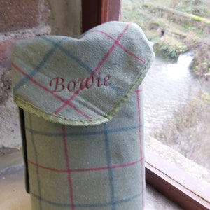 May include: A green plaid flask cover with red and blue lines, embroidered with the name "Bowie" in red. The cover has a flap with green trim and is made of a textured fabric.