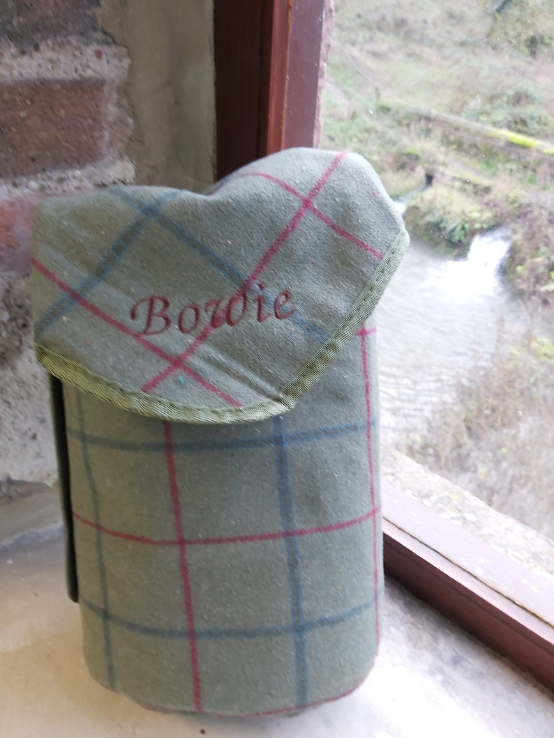 May include: A green plaid flask cover with red and blue lines, embroidered with the name "Bowie" in red. The cover has a flap with green trim and is made of a textured fabric.