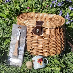 Può includere: Un cestino in vimini con chiusura in pelle, un coltello per funghi con spazzola, una piccola spazzola per funghi e una tazza con un'illustrazione di funghi, in un'area erbosa con fiori selvatici.