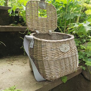 May include: A wicker basket with a light brown finish and a light gray strap. The basket has a hinged lid and a metal clasp. The basket is sitting on a set of stone steps.