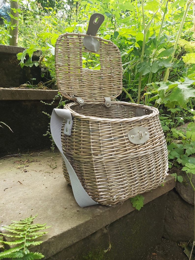 May include: A wicker basket with a light brown finish and a light gray strap. The basket has a hinged lid and a metal clasp. The basket is sitting on a set of stone steps.