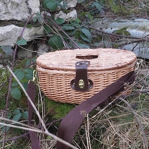 Może przedstawiać: Koszyk z wikliny w kolorze brązowym z brązowym paskiem skórzanym i złotym metalowym zapięciem. Koszyk leży na podłożu z zielonego mchu i brązowych gałązek.