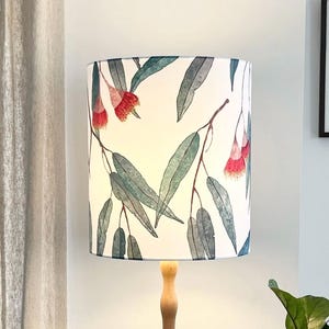May include: A tall wooden floor lamp with a cylindrical lampshade. The lampshade features a white background with a watercolor design of eucalyptus leaves and red flowers. The lamp has a light wood finish.