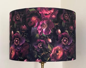 Può includere: Un paralume con una stampa floreale scura con fiori viola e rosa intenso su uno sfondo nero. Il paralume è realizzato in tessuto e ha una forma cilindrica.
