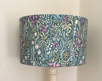May include: A table lamp with a cylindrical drum shade featuring a teal background with a dense botanical pattern of purple, white, and light green flowers and foliage. The lamp base is a cream-coloured, fluted column with a circular pedestal foot.