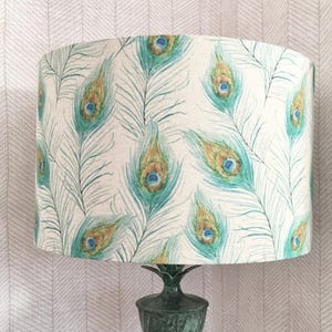 May include: A white lampshade featuring a peacock feather design in shades of teal, green, and gold. The lampshade is cylindrical and has a smooth, fabric texture.