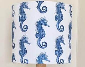 May include: A wooden table lamp with a white lampshade featuring a repeating pattern of blue seahorses. The lamp has a conical wooden base and is placed on a light-colored wooden surface.