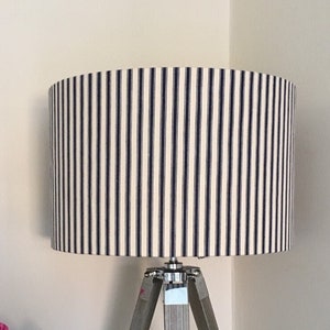 May include: A table lamp with a cylindrical lampshade featuring a navy blue and white vertical pinstripe pattern. The lamp base consists of three light grey wooden legs arranged in a tripod configuration with silver metal hardware.