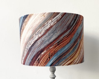 May include: A lampshade with a cylindrical shape, featuring an abstract design of swirling lines in brown, blue, grey, and white. It is mounted on a grey lamp stand, forming a decorative lighting fixture.