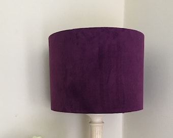 May include: A white table lamp with a purple velvet lampshade. The lamp is on a wooden table with a vase of white roses.