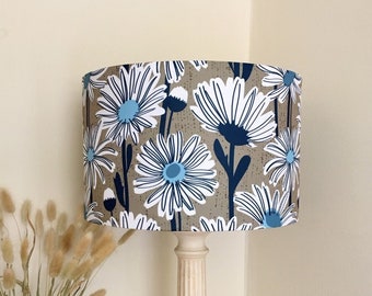 May include: A white table lamp with a beige and blue floral patterned lampshade. The lampshade has a white background with blue and white flowers and dark blue stems. The lamp base is white and has a fluted design.