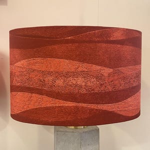 May include: A cylindrical lamp shade featuring a textured, wavy pattern in shades of deep red and burnt orange. The surface has a distinct, tactile finish with horizontal bands of varying red tones that create a layered, undulating visual effect.
