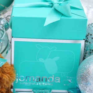 May include: A turquoise blue gift box with a white ribbon and bow. The box has a silver trim and the words "jomanda" and "Softer than a soft thing" printed on the front.