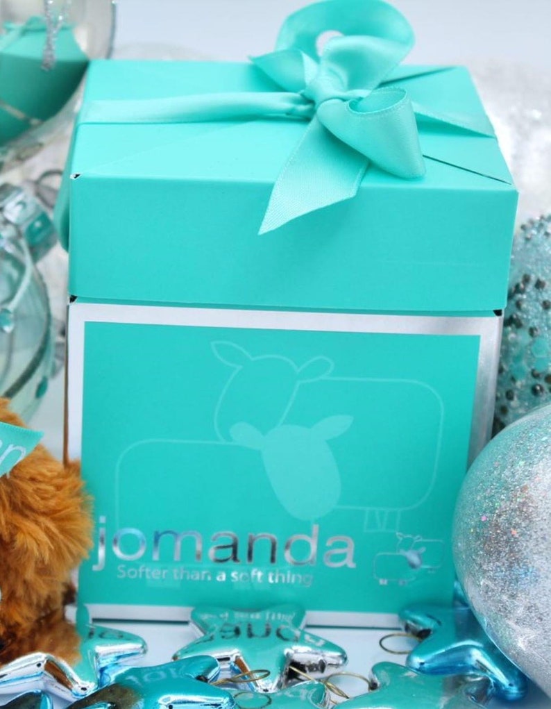 May include: A turquoise blue gift box with a white ribbon and bow. The box has a silver trim and the words "jomanda" and "Softer than a soft thing" printed on the front.
