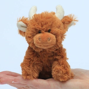 May include: A small, brown plush Highland cow toy with white horns and ears. The toy is sitting on a person's hand.