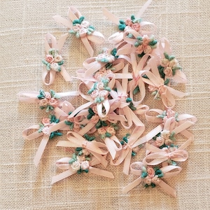 May include: A pile of pink satin ribbon bows with green and pink floral embroidery. The bows are scattered on a beige fabric background.