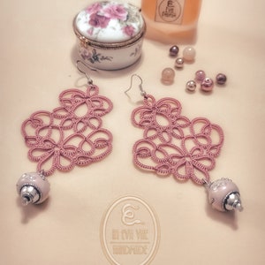 May include: A pair of pink crocheted earrings with a pearl-like bead on each earring. The earrings are hanging from silver hooks. The earrings are on a light pink background with a small porcelain box with pink roses and a bottle of perfume.