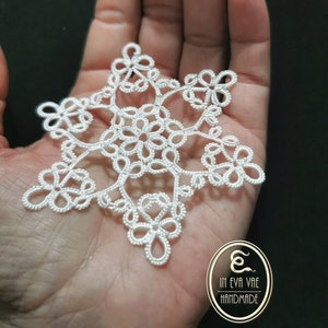 Può includere: Un delicato fiocco di neve bianco fatto a crochet. Il fiocco di neve è intricato e dettagliato, con una forma a stella a sei punte. Il fiocco di neve è tenuto in una mano, e lo sfondo è nero.
