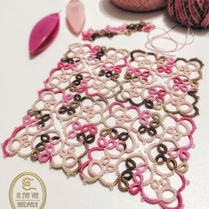 May include: A delicate, intricate lace doily in shades of pink, brown, and beige. The doily is handmade and features a repeating pattern of flowers and leaves. The doily is laid out on a white surface.