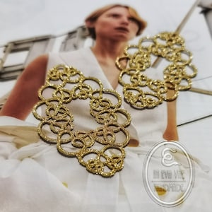 Può includere: Orecchini dorati a cerchi aperti dall'aspetto strutturato e glitterato. Gli orecchini sono disposti su una superficie bianca, con uno sfondo sfocato con una persona con un top bianco. Fatto a mano.