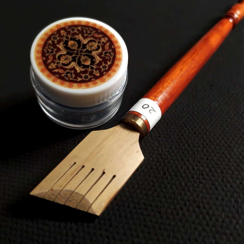 May include: A wooden calligraphy pen with a brown handle and a brass tip. The pen has a white label with the number "20" on it. The pen is next to a small clear jar with a lid that has a colourful patterned design.