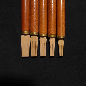 May include: Five calligraphy pens with cylindrical wooden handles and brass ferrules. Each pen features a flat, light-coloured bamboo nib with a central slit, arranged in a row from widest to narrowest tip, showcasing natural wood grain textures.