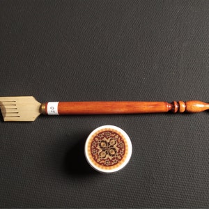 May include: A wooden calligraphy pen with a brown handle and a gold metal nib. The pen has a small, round, decorative design on the handle. The pen is next to a small, round, white container with a brown and gold design.