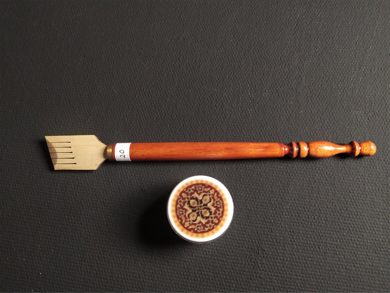 May include: A wooden calligraphy pen with a brown handle and a gold metal nib. The pen has a small, round, decorative design on the handle. The pen is next to a small, round, white container with a brown and gold design.