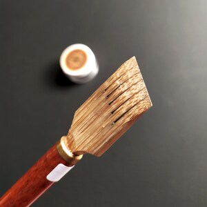 May include: A wooden calligraphy brush with a brown handle and a light brown bamboo brush head with five slits. The brush is on a black background.
