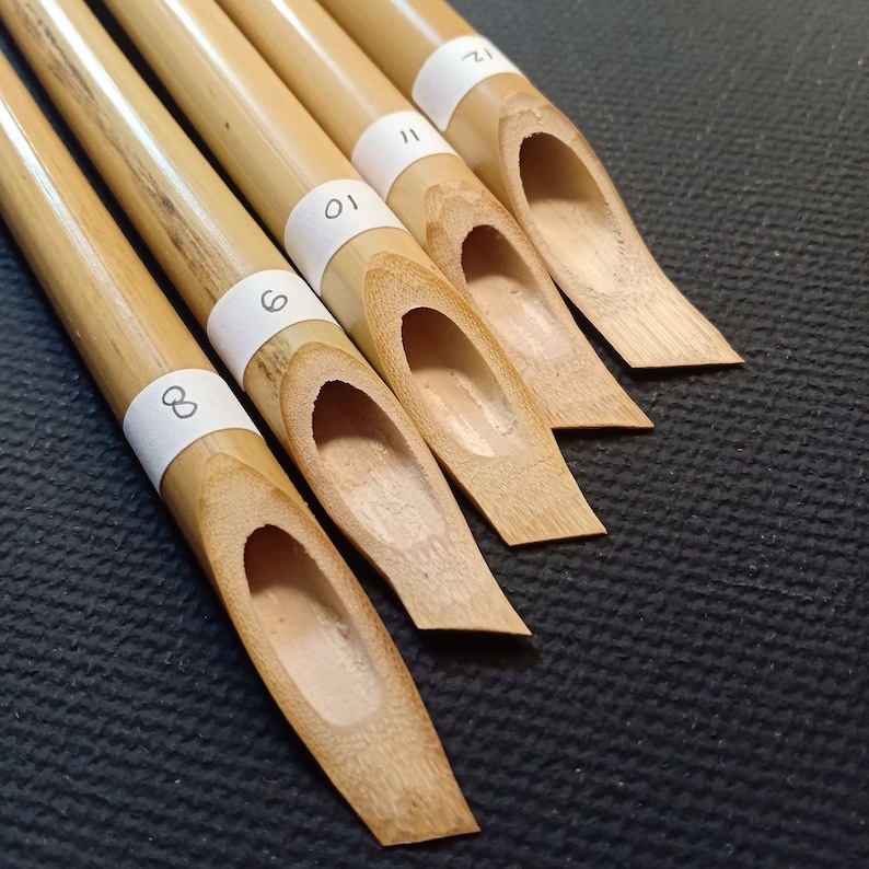 May include: Five bamboo calligraphy pens with numbered labels. The pens are arranged in a fan shape, with the largest pen at the bottom and the smallest pen at the top. The pens are all natural bamboo color.