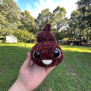 May include: A plush, burgundy-colored crocheted poop emoji with large, glossy black eyes and a white smiling mouth. The whimsical design is held against a backdrop of green grass and trees, suggesting an outdoor setting.