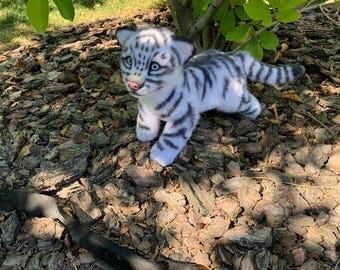 May include: A plush toy tiger cub with white fur and black stripes. The stuffed animal features bright blue eyes, a small pink nose, and a long striped tail. It is standing on all four paws, with a realistic facial expression and soft textured fur.