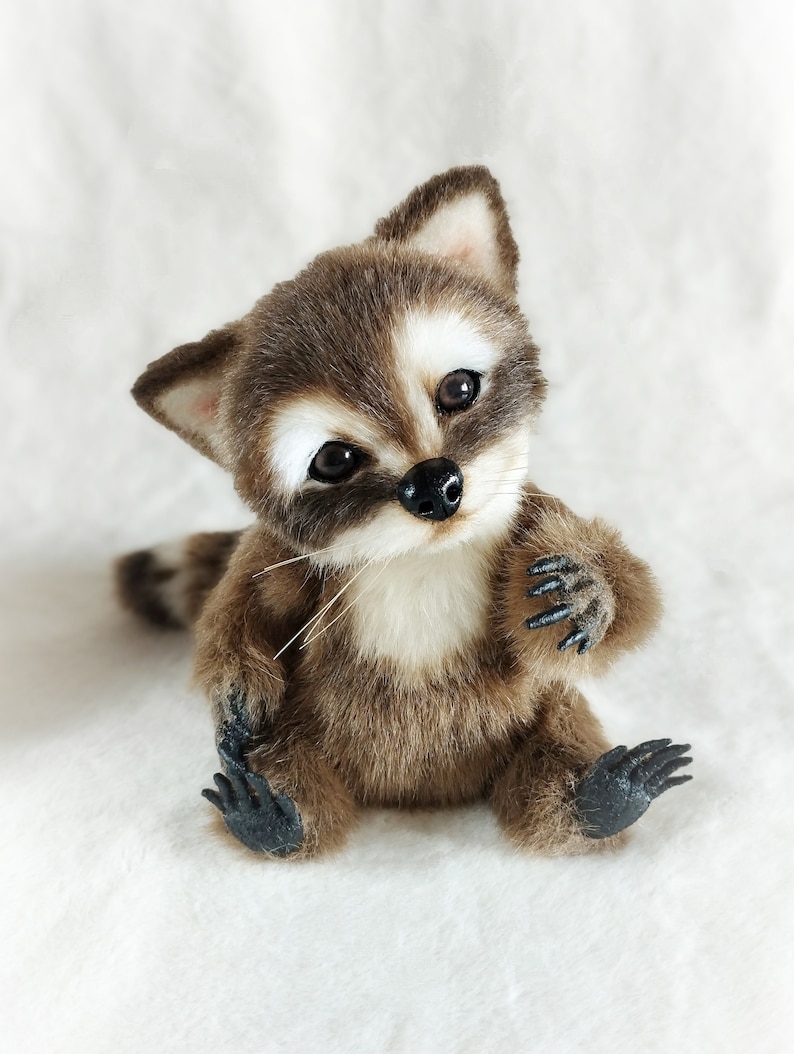 May include: A small, brown and white plush raccoon toy with black eyes and a black nose. The raccoon is sitting up with its paws in front of it. It has a white chest and a black tail.