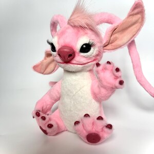 Puede incluir: Un peluche rosa con orejas grandes, pesta&ntilde;as negras y un mech&oacute;n de pelo rosa. El juguete tiene una barriga blanca y patas rosas con almohadillas de color rojo oscuro. Est&aacute; sobre una superficie blanca.