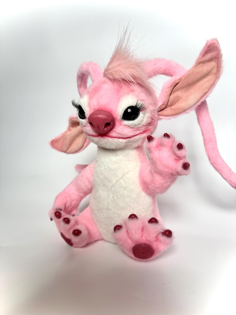 Puede incluir: Un peluche rosa con orejas grandes, pesta&ntilde;as negras y un mech&oacute;n de pelo rosa. El juguete tiene una barriga blanca y patas rosas con almohadillas de color rojo oscuro. Est&aacute; sobre una superficie blanca.