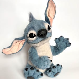 Puede incluir: Un personaje de peluche azul y blanco con grandes ojos negros, una nariz negra y orejas grandes. El personaje tiene una expresi&oacute;n amigable y est&aacute; sentado con una pata levantada. El juguete de peluche tiene una textura suave y peluda.