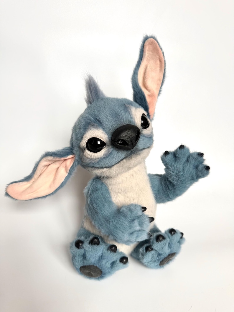Puede incluir: Un personaje de peluche azul y blanco con grandes ojos negros, una nariz negra y orejas grandes. El personaje tiene una expresi&oacute;n amigable y est&aacute; sentado con una pata levantada. El juguete de peluche tiene una textura suave y peluda.