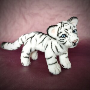 Op de afbeelding: Een wit knuffeltijgerwelpje met zwarte strepen. De tijger heeft blauwe ogen en staat op een roze oppervlak.