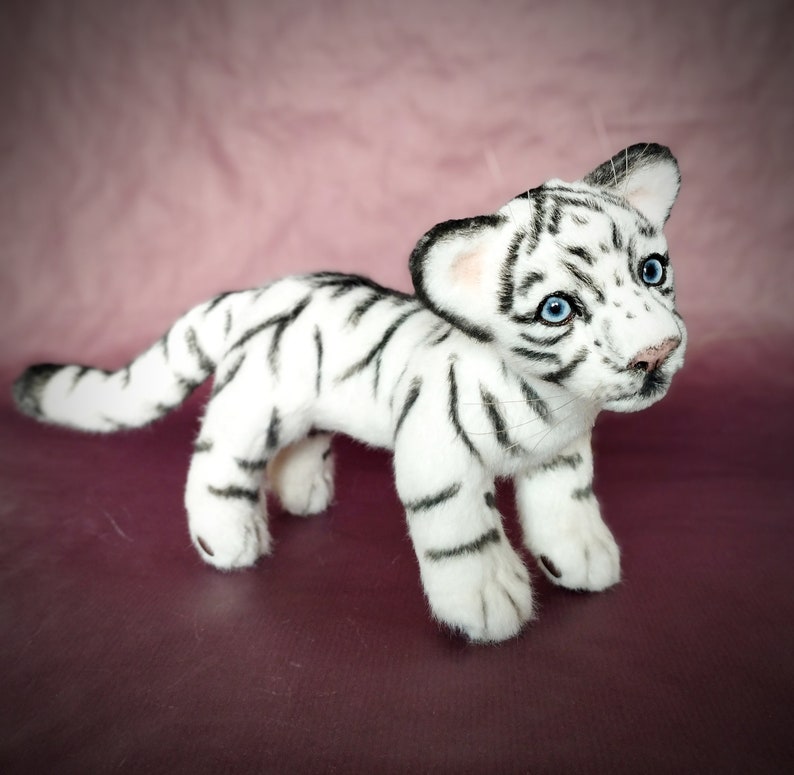 Op de afbeelding: Een wit knuffeltijgerwelpje met zwarte strepen. De tijger heeft blauwe ogen en staat op een roze oppervlak.