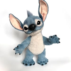 Puede incluir: Un personaje de peluche azul y blanco con orejas grandes, ojos negros y una nariz negra. El personaje tiene una expresi&oacute;n amigable y est&aacute; de pie con los brazos extendidos. El juguete de peluche tiene una textura suave y peluda.