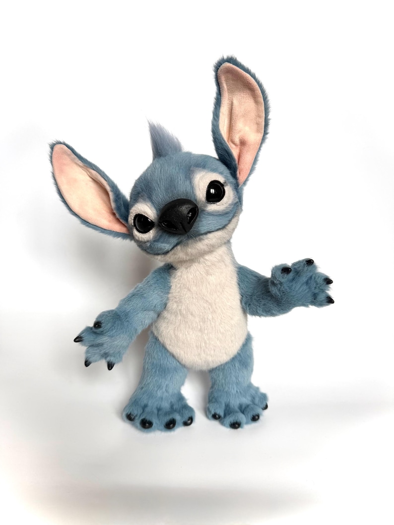 Puede incluir: Un personaje de peluche azul y blanco con orejas grandes, ojos negros y una nariz negra. El personaje tiene una expresi&oacute;n amigable y est&aacute; de pie con los brazos extendidos. El juguete de peluche tiene una textura suave y peluda.