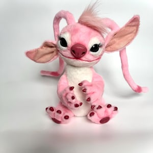 Puede incluir: Una criatura de peluche rosa con orejas grandes, antenas largas y un mech&oacute;n de pelo. Tiene ojos negros con largas pesta&ntilde;as, una nariz rosa y garras rojas. La criatura est&aacute; sentada sobre una superficie blanca.