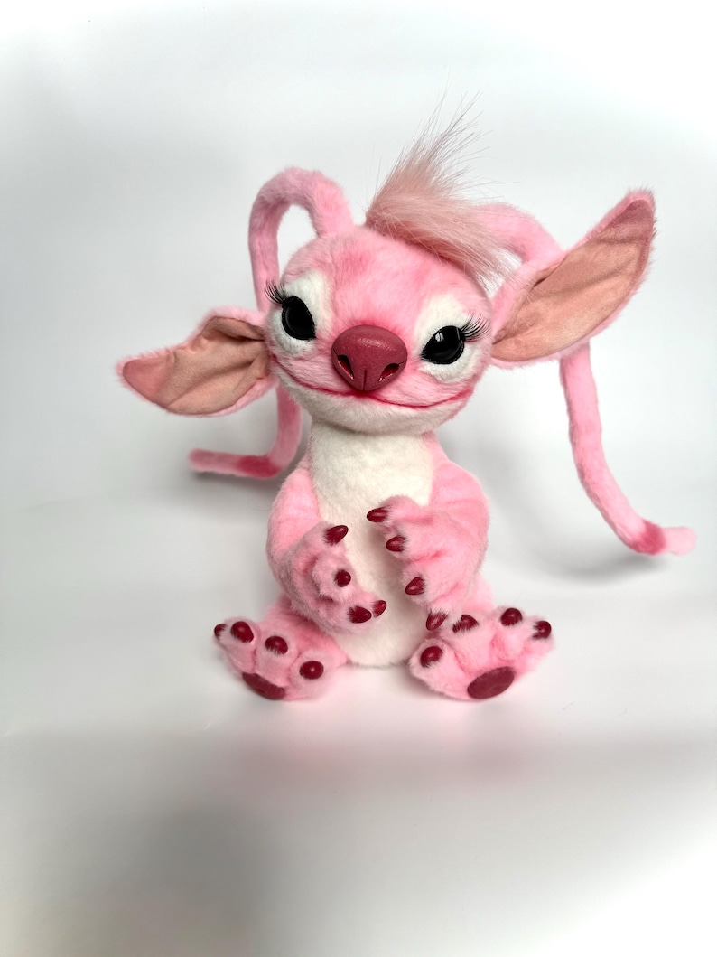 Puede incluir: Una criatura de peluche rosa con orejas grandes, antenas largas y un mech&oacute;n de pelo. Tiene ojos negros con largas pesta&ntilde;as, una nariz rosa y garras rojas. La criatura est&aacute; sentada sobre una superficie blanca.
