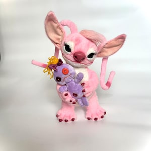 May include: A plush toy character with light pink fur, large ears, and long eyelashes, holding a smaller purple doll. The purple doll features yellow yarn hair, a dark purple bow, a single orange button eye, and stitched mouth details.
